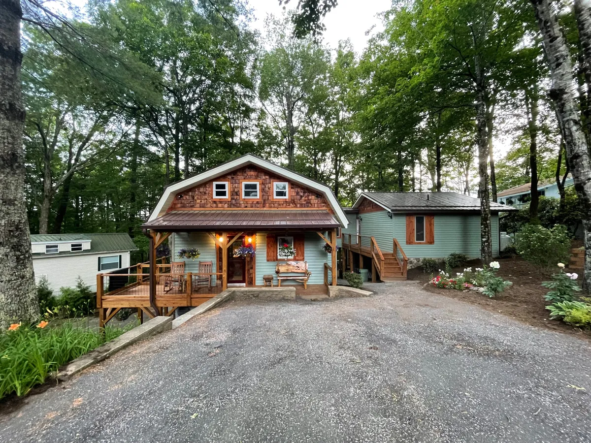 The image features a beautifully maintained exterior of a single-level mountain home with a spacious wooden deck, inviting front steps, lush landscaped garden, and large windows that let in natural light. The cozy deck space is perfect for gatherings and relaxation, all set against a tranquil, wooded backdrop.
