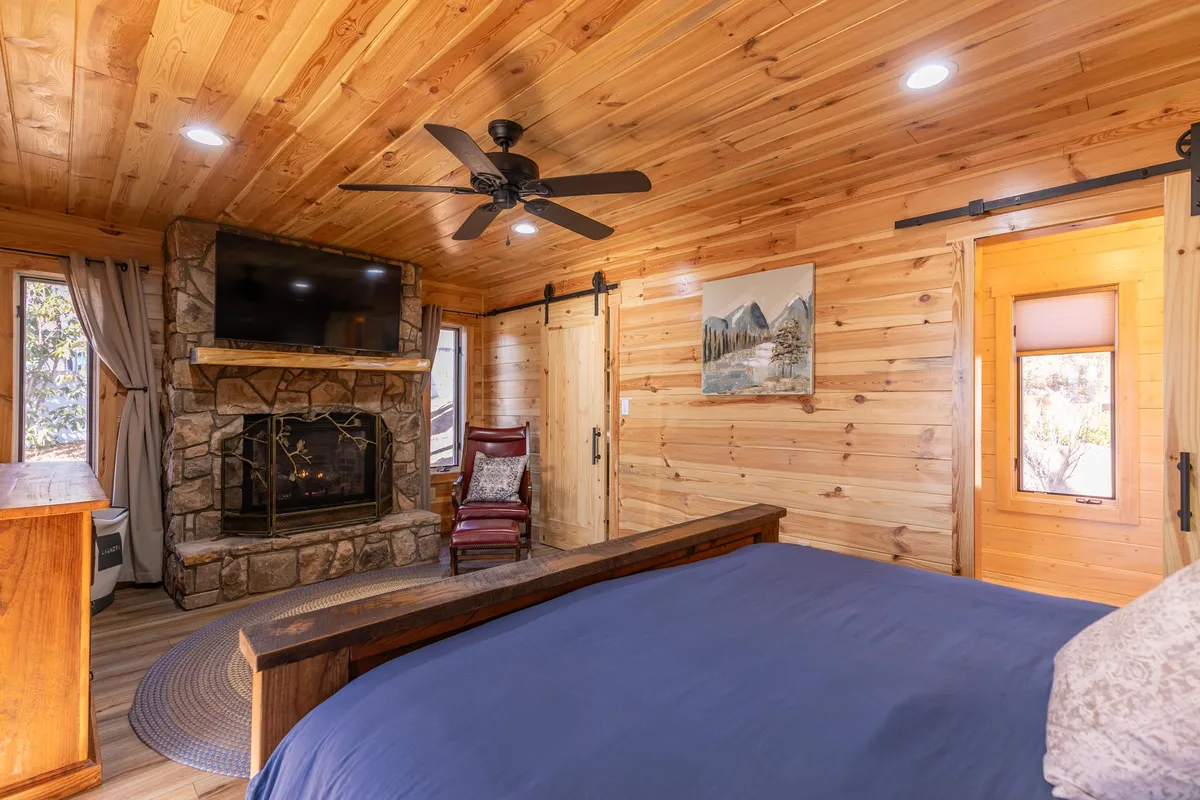 A spacious primary bedroom featuring a wooden bed with plush bedding, a prominent stone fireplace, a ceiling fan, and a large window looking out into nature. The room exudes warmth with its inviting textures and natural materials, perfect for unwinding after a day outdoors.