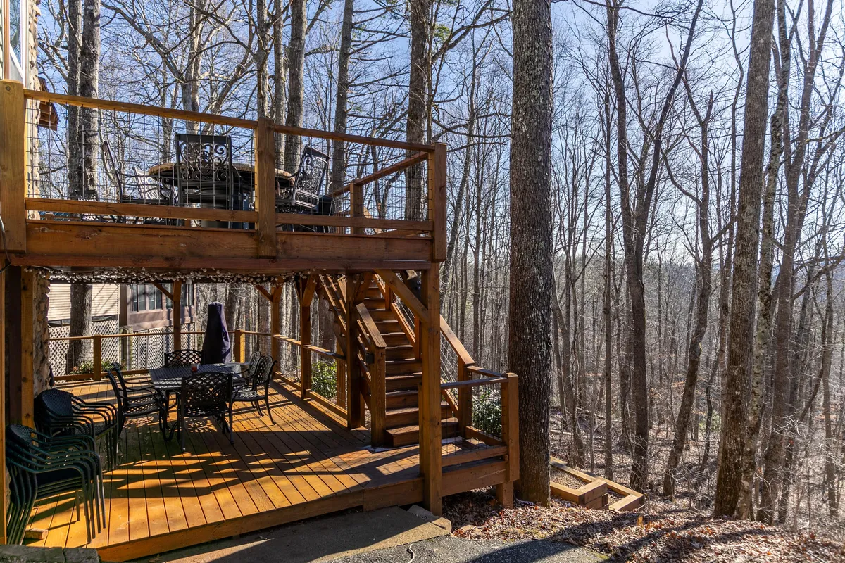 A wooden deck surrounded by trees, featuring black outdoor furniture and stairs leading up. The space exudes tranquility, perfect for outdoor dining or relaxation while being immersed in peaceful wooded surroundings.