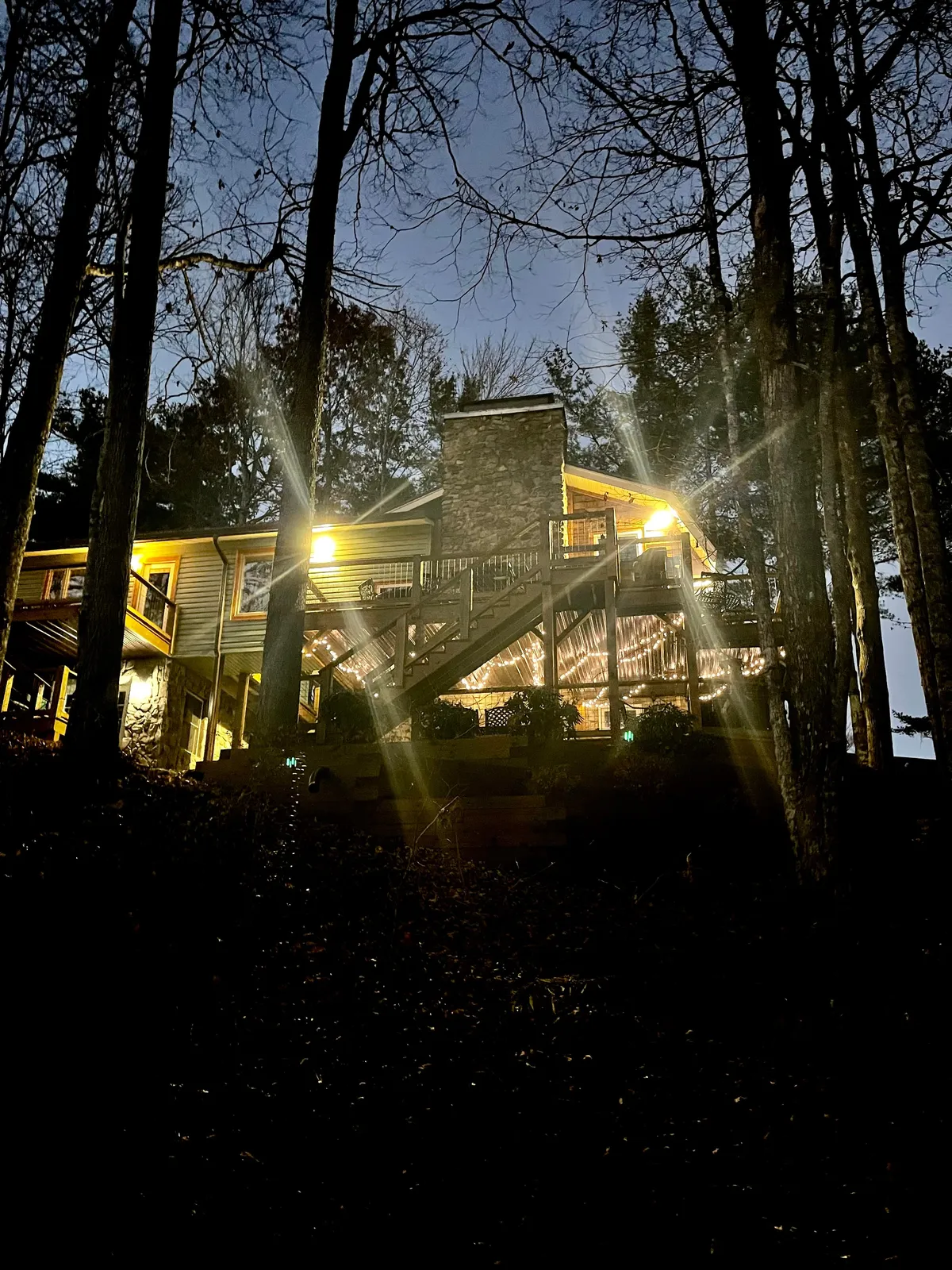 This warm and welcoming evening exterior highlights the illuminated house nestled among trees, with a fire pit area glowing in the foreground. The setting suggests gatherings around the fire, storytelling, or making s’mores, all framed by the peacefulness of nature at dusk.