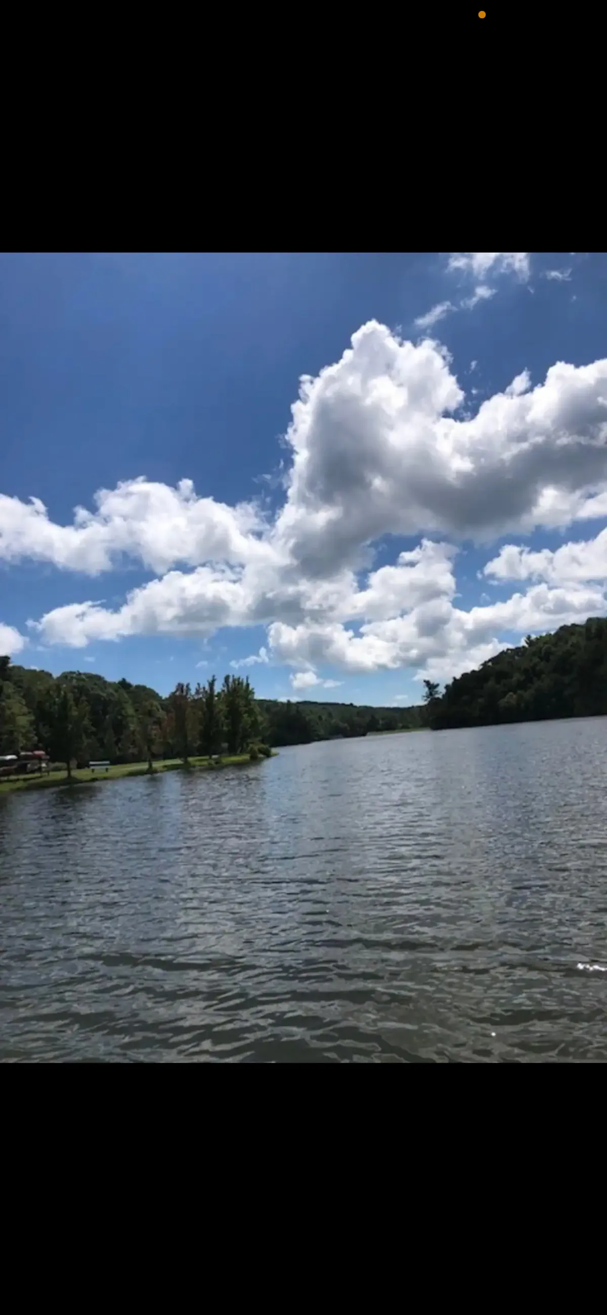 A broad view across a glassy lake with a distant tree line and fluffy clouds overhead. The tranquil water invites boating, fishing, or simply relaxing by the shore while taking in the scenery.