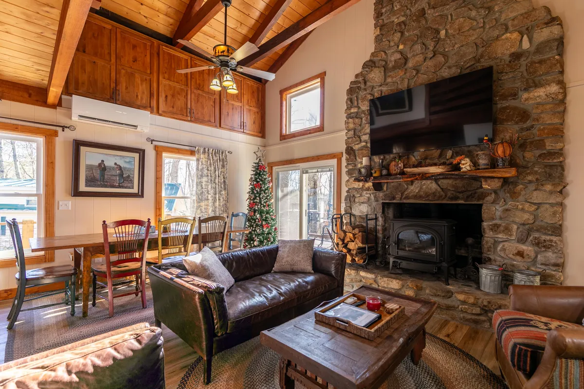 A warm living area with a striking stone fireplace, large windows framing scenic views, a festive Christmas tree, and a wooden dining table. The ambiance is both homey and celebratory, with natural light flooding the space and a clear view into the dining area, underscoring the communal spirit of the home.