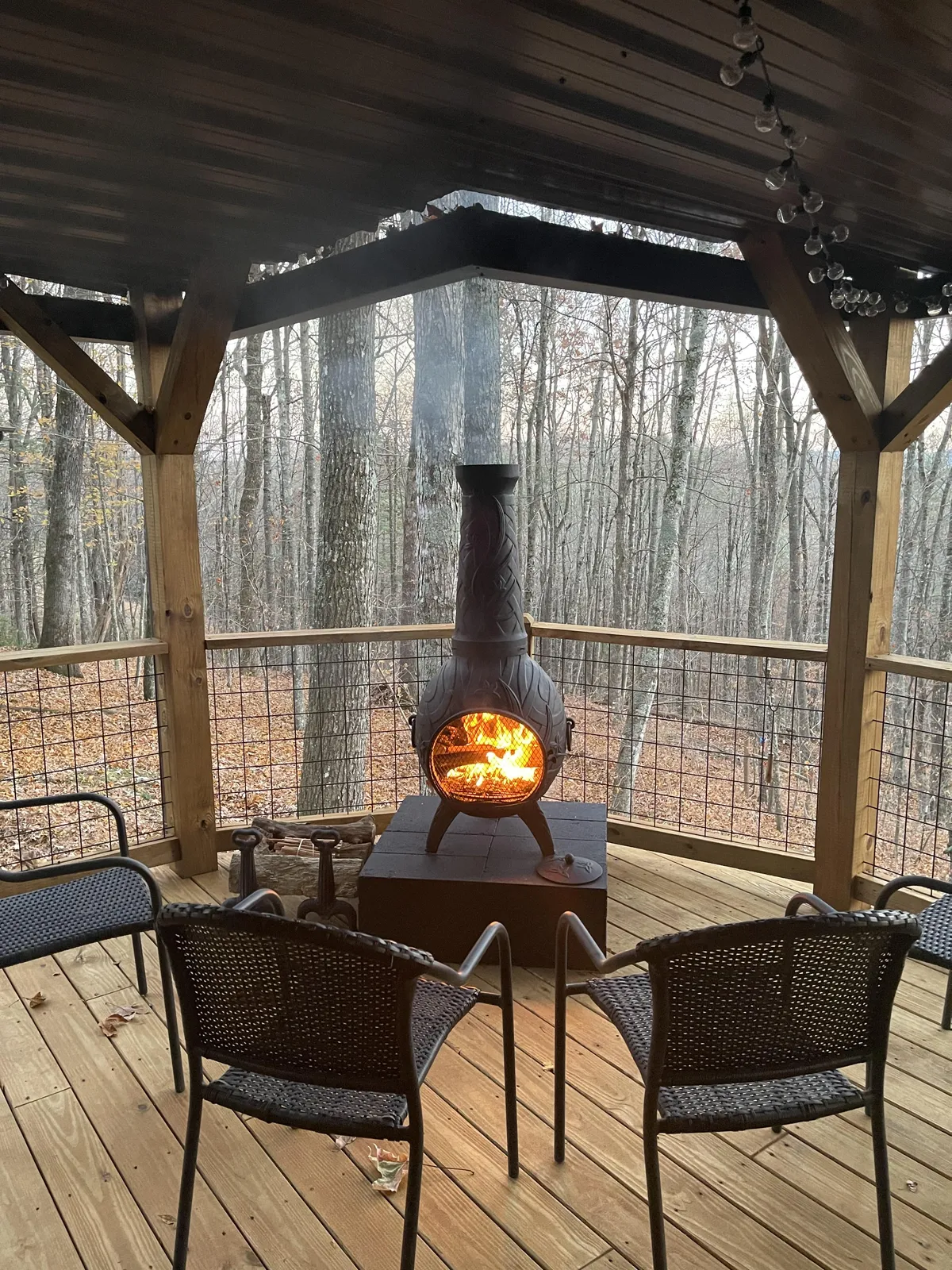 A warm fire table is surrounded by comfortable deck chairs on a wooden deck, with trees in the background and a cozy, inviting atmosphere. The image captures the ambiance of gathering with friends or family, sharing stories by the fire, and enjoying the outdoors.