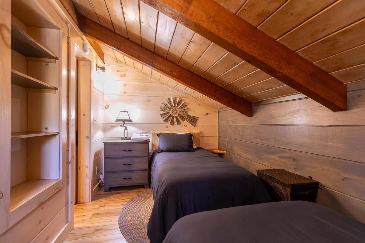 A cheerful fourth bedroom with two twin beds, a central nightstand, a lamp, and decorative wall art. Natural light filters across the wooden beams and soft bedding, creating a welcoming space for family or friends.