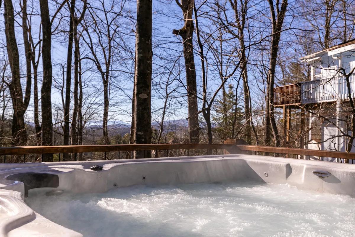 This image shows a hot tub nestled on a wooden deck, surrounded by towering trees and distant mountain views. The setting is tranquil and immersive, suggesting privacy and a strong connection to nature. At night, you can imagine the stars overhead, while in the morning, sunlight fills the deck.