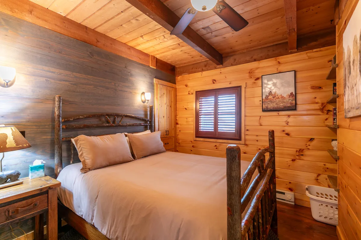 A cozy guest bedroom with a wooden bed frame, rustic wooden walls, soft lighting, and tasteful artwork. Sunlight streams through a shuttered window, creating a tranquil and intimate ambiance ideal for restful nights.